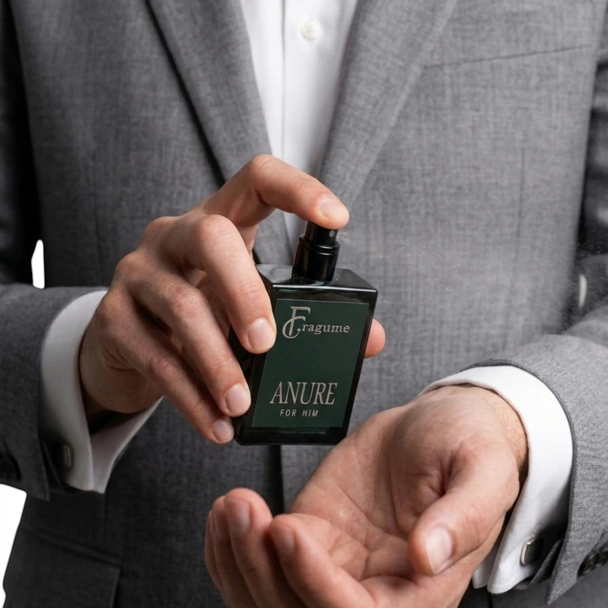 Man in a grey suit spraying Fragume Anure Parfum from a sleek forest green glass bottle onto his wrist.